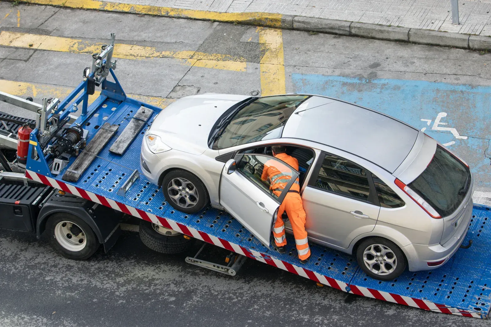 Tow truck loading a silver car from a disabled parking spot. A person in orange is exiting the car.