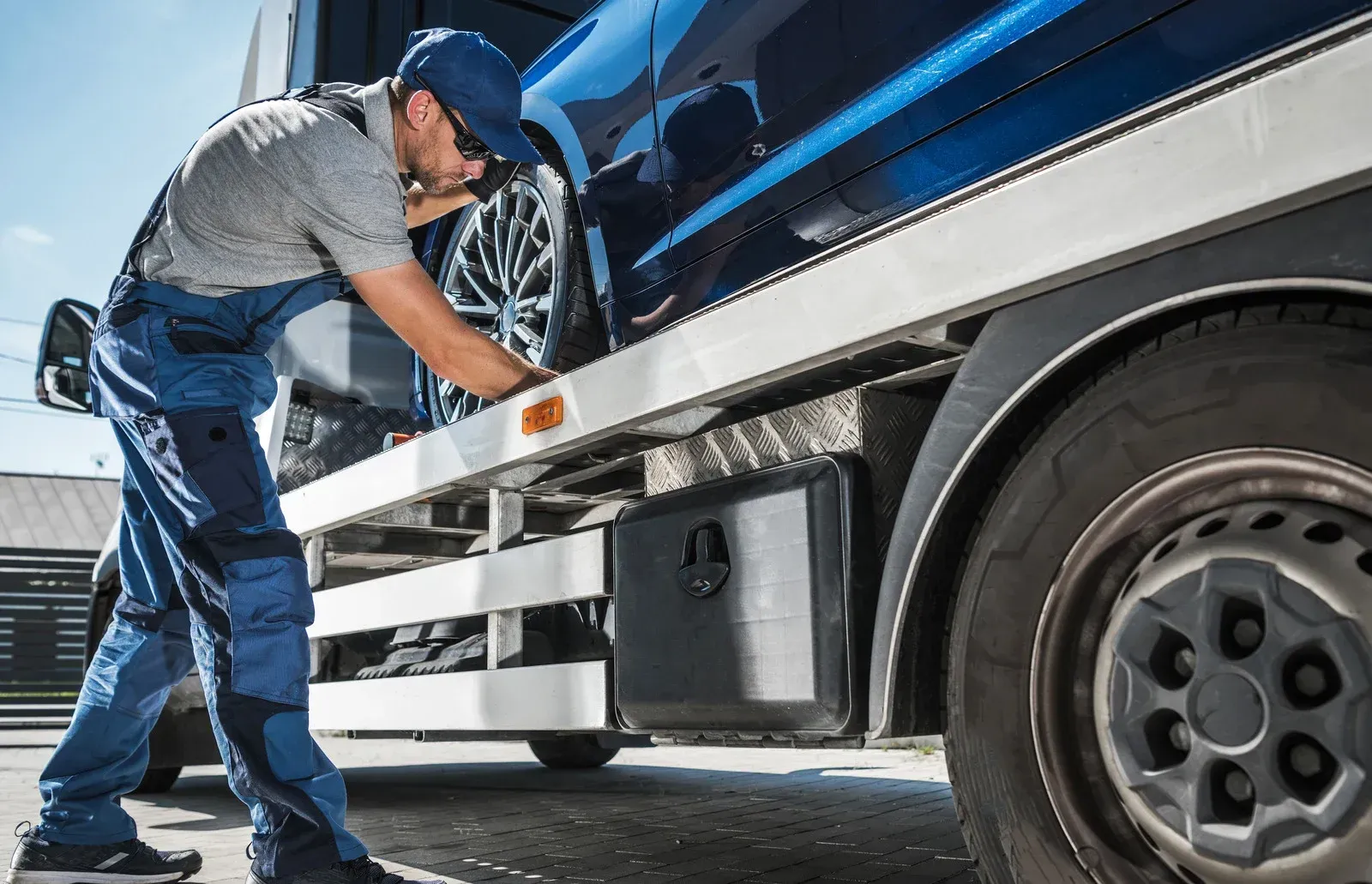 Mechanic in blue overalls inspecting the underside of a blue and silver truck near a tire.