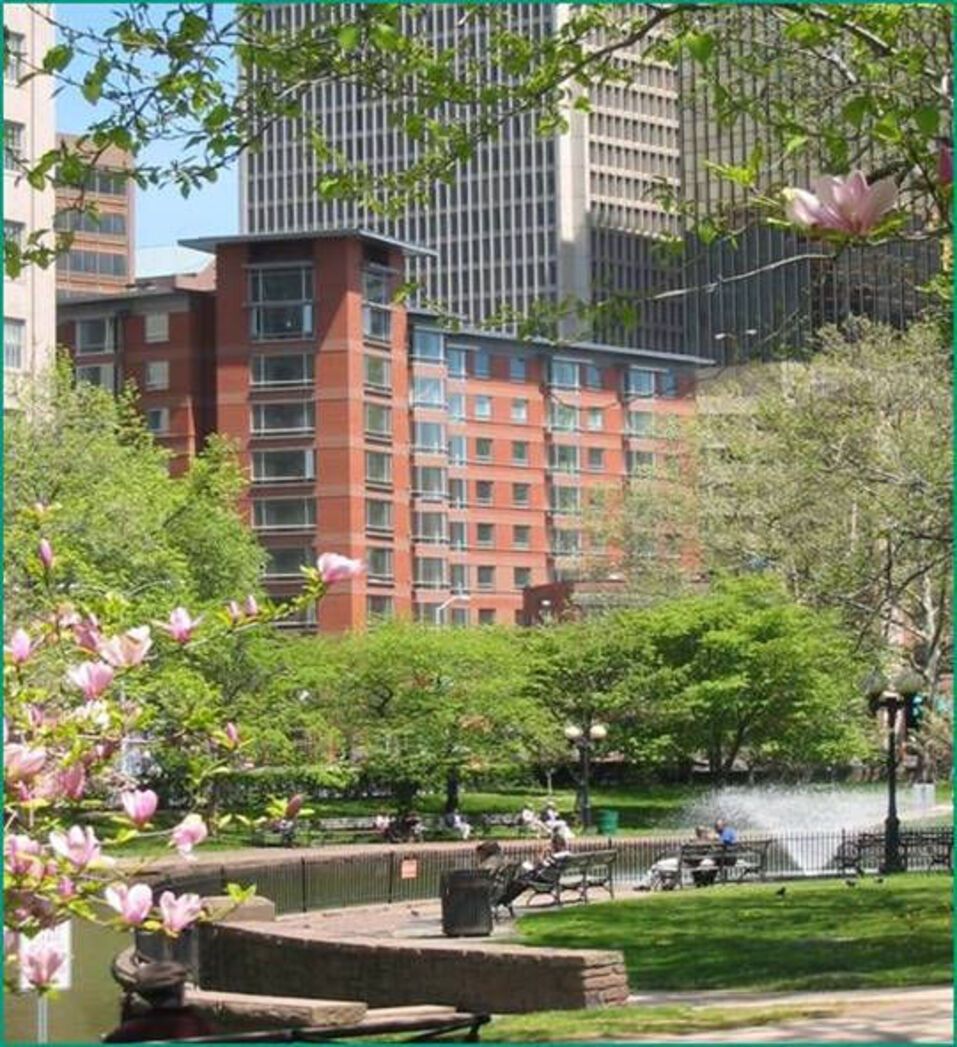Brick apartment building in a park with a fountain, trees with pink blossoms, and tall buildings in the background.