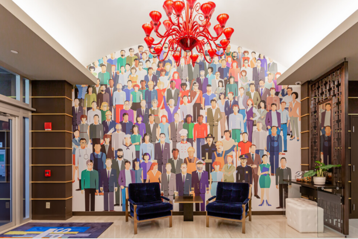 A hotel lobby with a mural of diverse people, two blue velvet chairs, and a red chandelier.