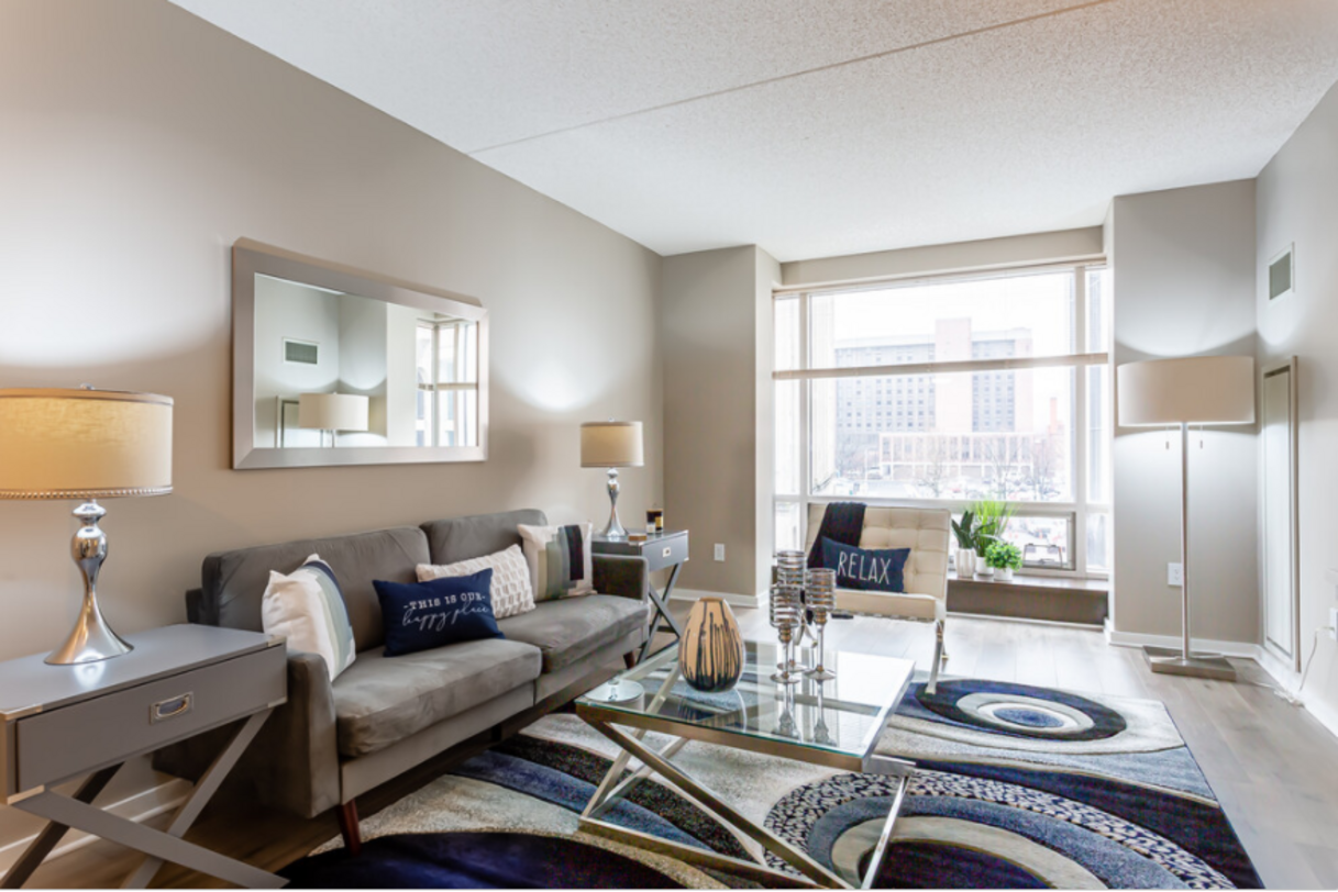 Living room with a gray sofa, side tables, and a large window overlooking city buildings. Features a blue and white rug.