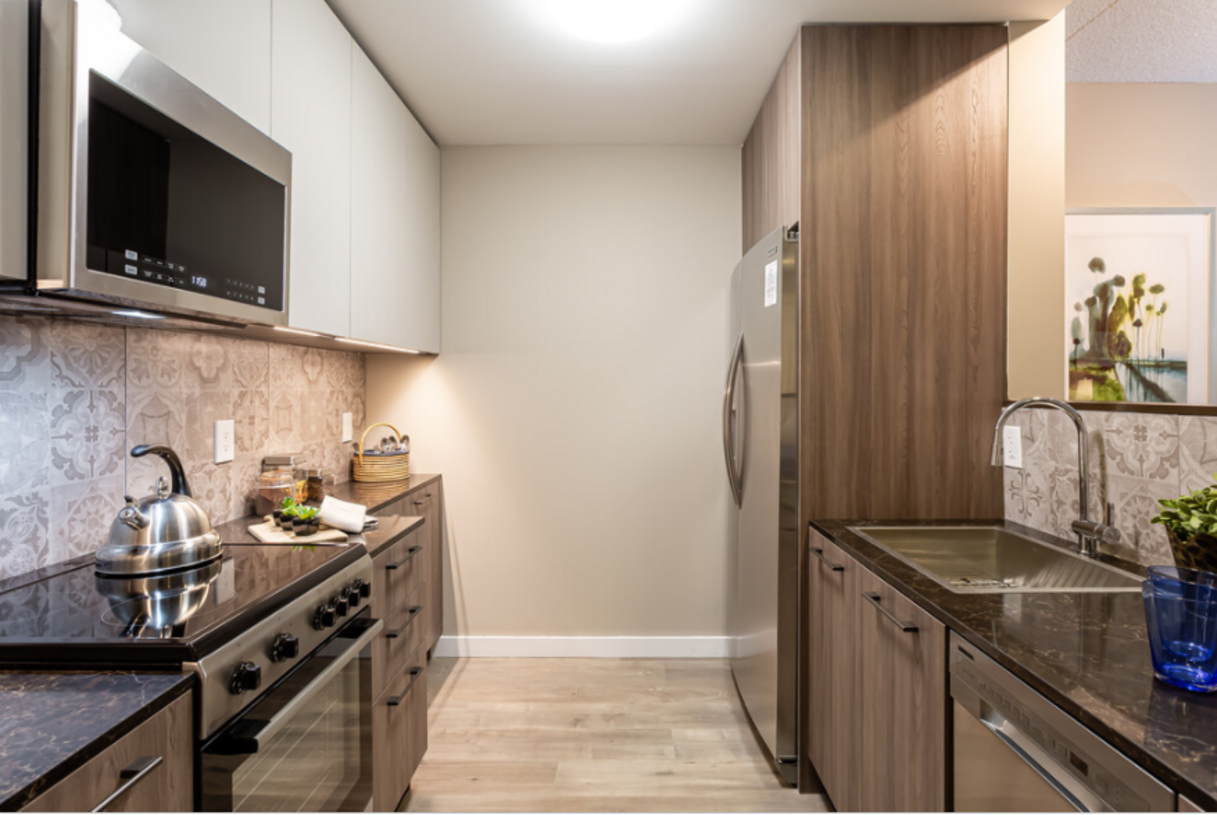 A narrow modern kitchen with wood-grain cabinets, stainless steel appliances, and black countertops.