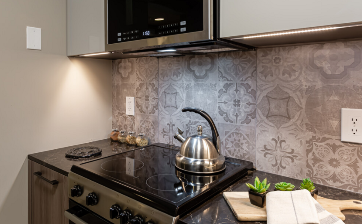 Kitchen with a stovetop, microwave, and decorative backsplash. A stainless steel kettle sits on the cooktop.