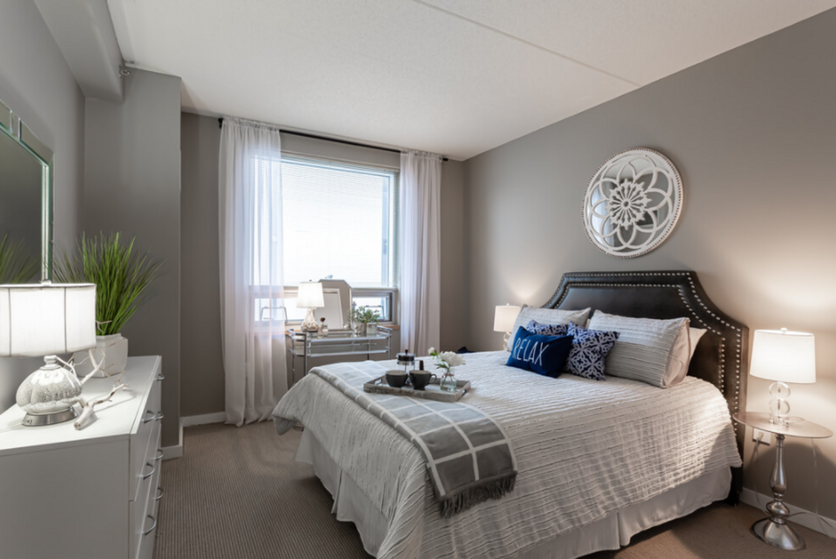 A bedroom with a bed, dresser, and window. The walls are gray, with white bedding, and a decorative wall hanging.
