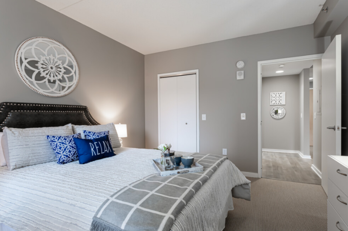 Cozy gray bedroom with a bed, decorative pillows, and a wall-mounted art piece. An open door leads to a hallway.