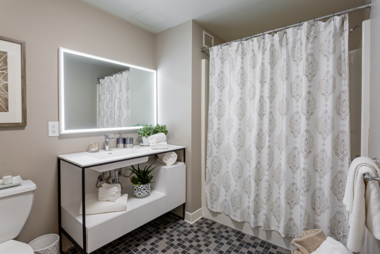 Modern bathroom with white vanity, illuminated mirror, patterned shower curtain, and patterned floor tiles.