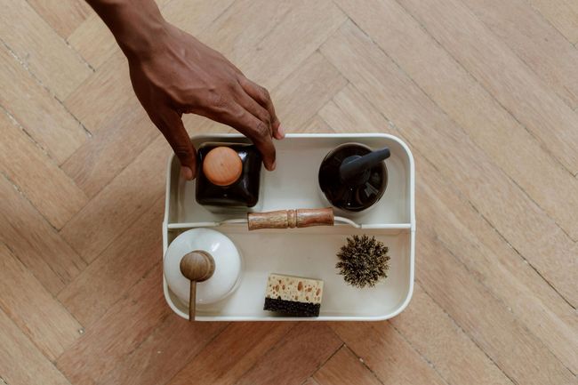 Hand placing a small cup on a white tray with tea utensils and a tea ball on a wooden floor