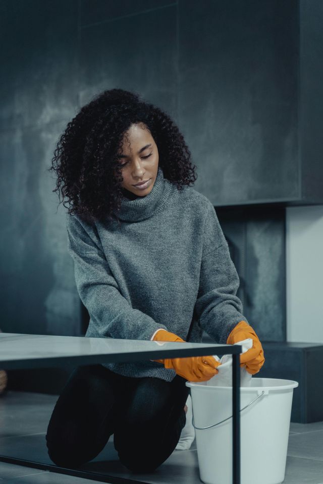 Person in gray sweater with orange gloves cleaning a table near a white bucket and cloth