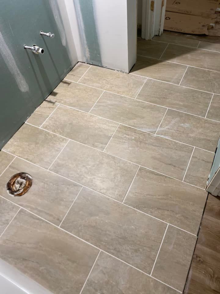 Bathroom floor with tan rectangular tiles and white grout; plumbing and walls visible.