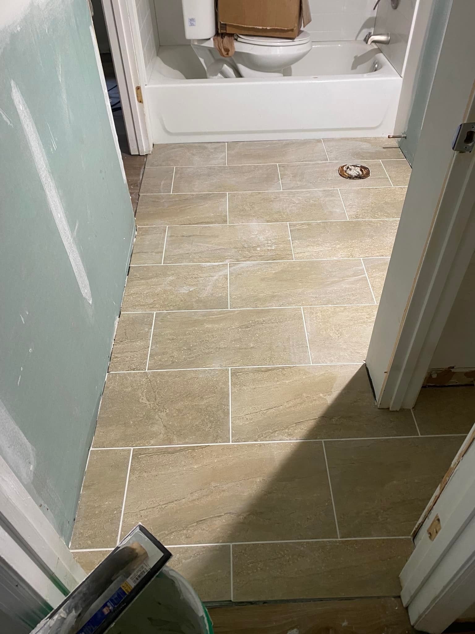 Bathroom floor tiling in progress, showing rectangular tiles, grout lines, and a toilet in the background.