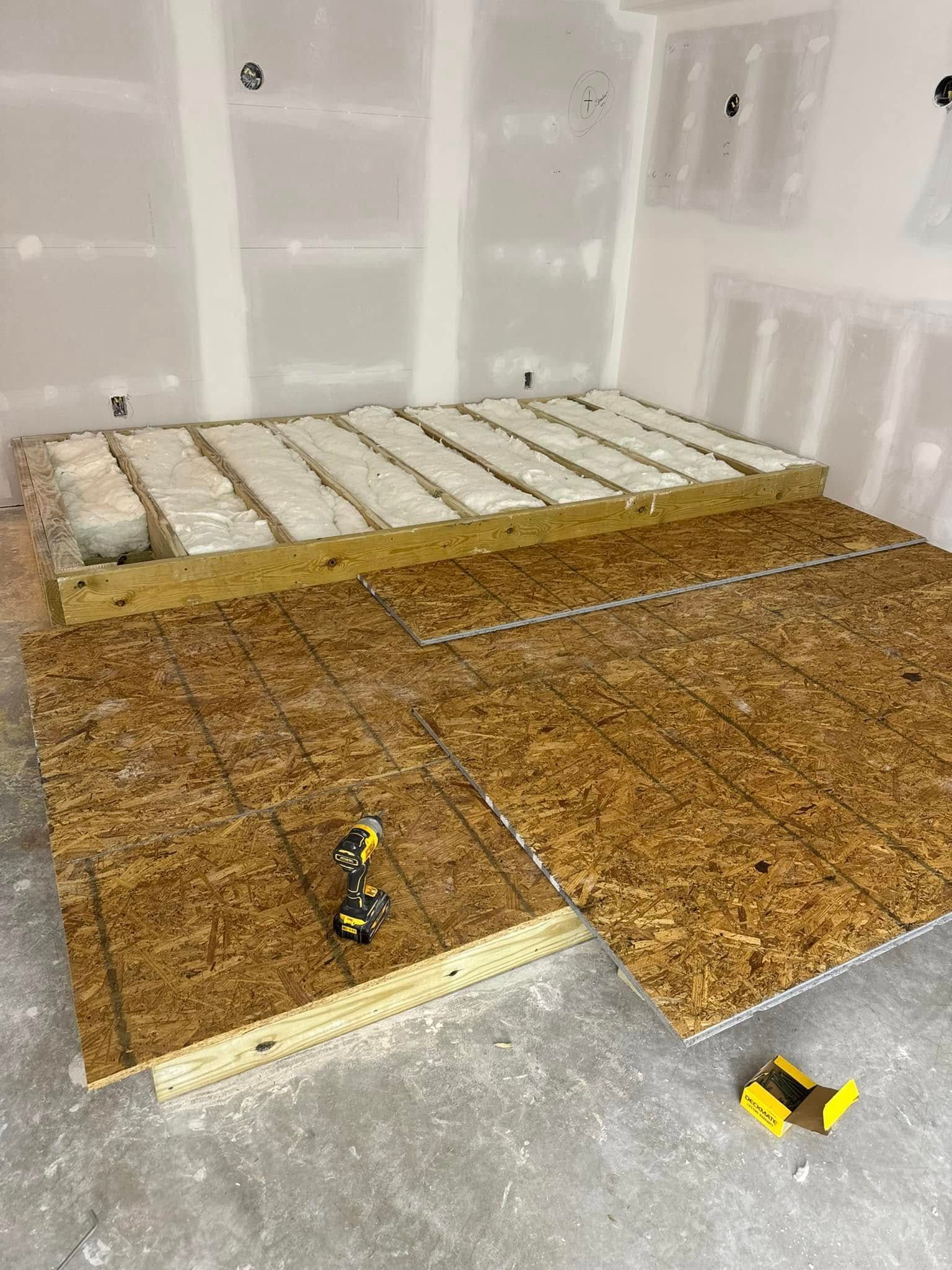 Construction site with plywood flooring being installed. Yellow tools visible. Walls are drywall.