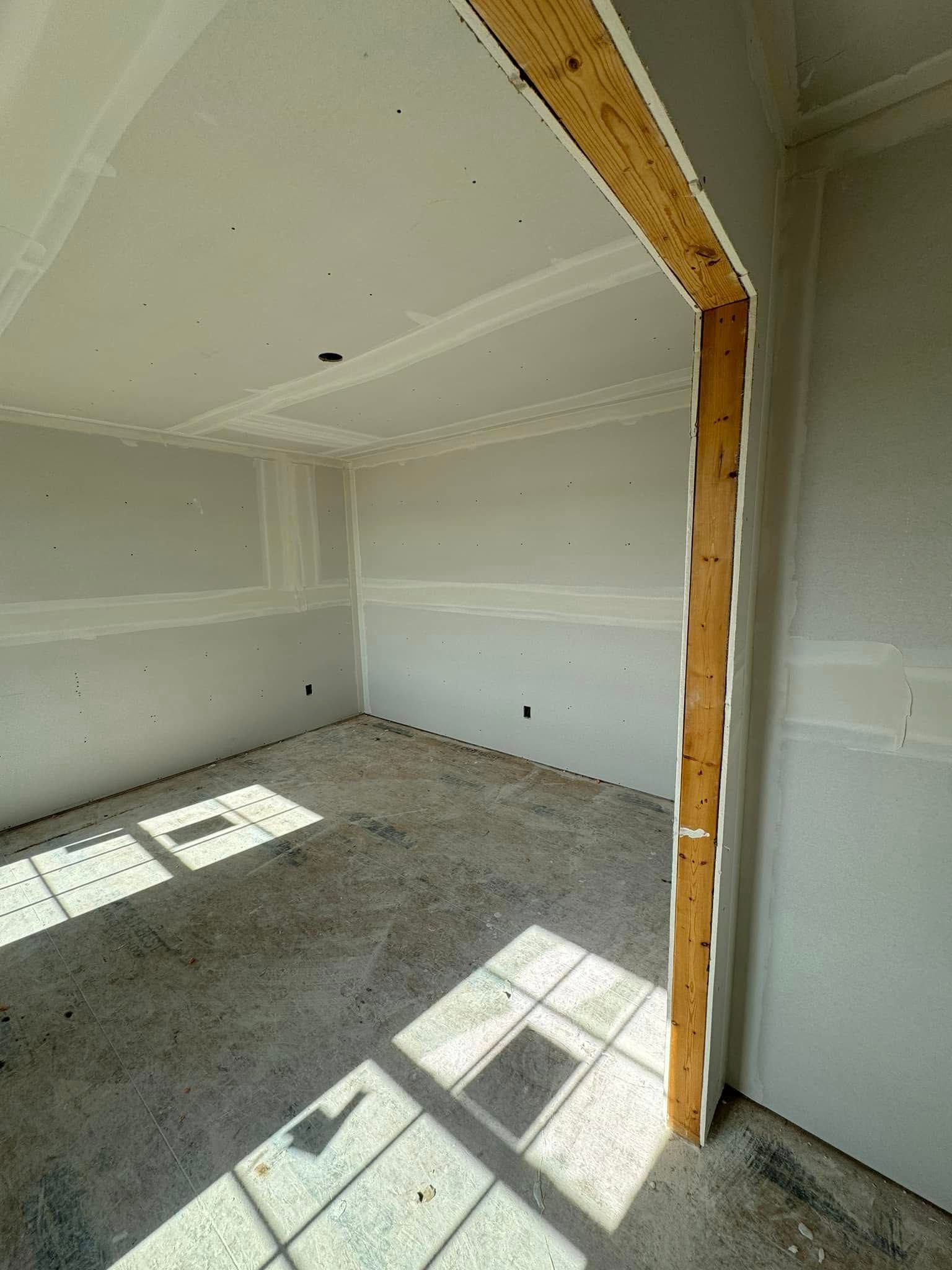 Interior room under construction, with drywall, unfinished flooring, and doorway framing. Sunlight creates shadows.
