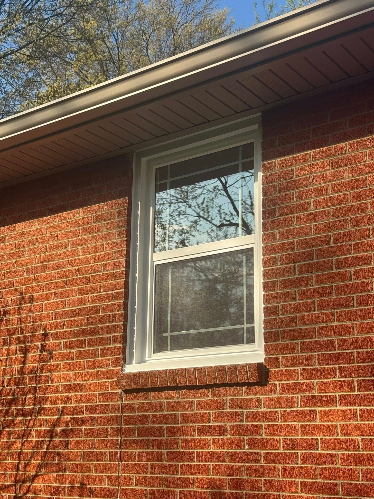 White framed window on red brick wall, under a brown roof with foliage reflection.