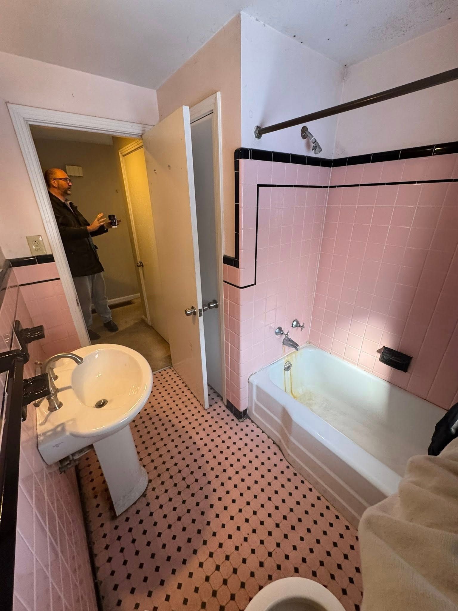 Pink-tiled bathroom with pedestal sink, bathtub, and a person in the doorway.
