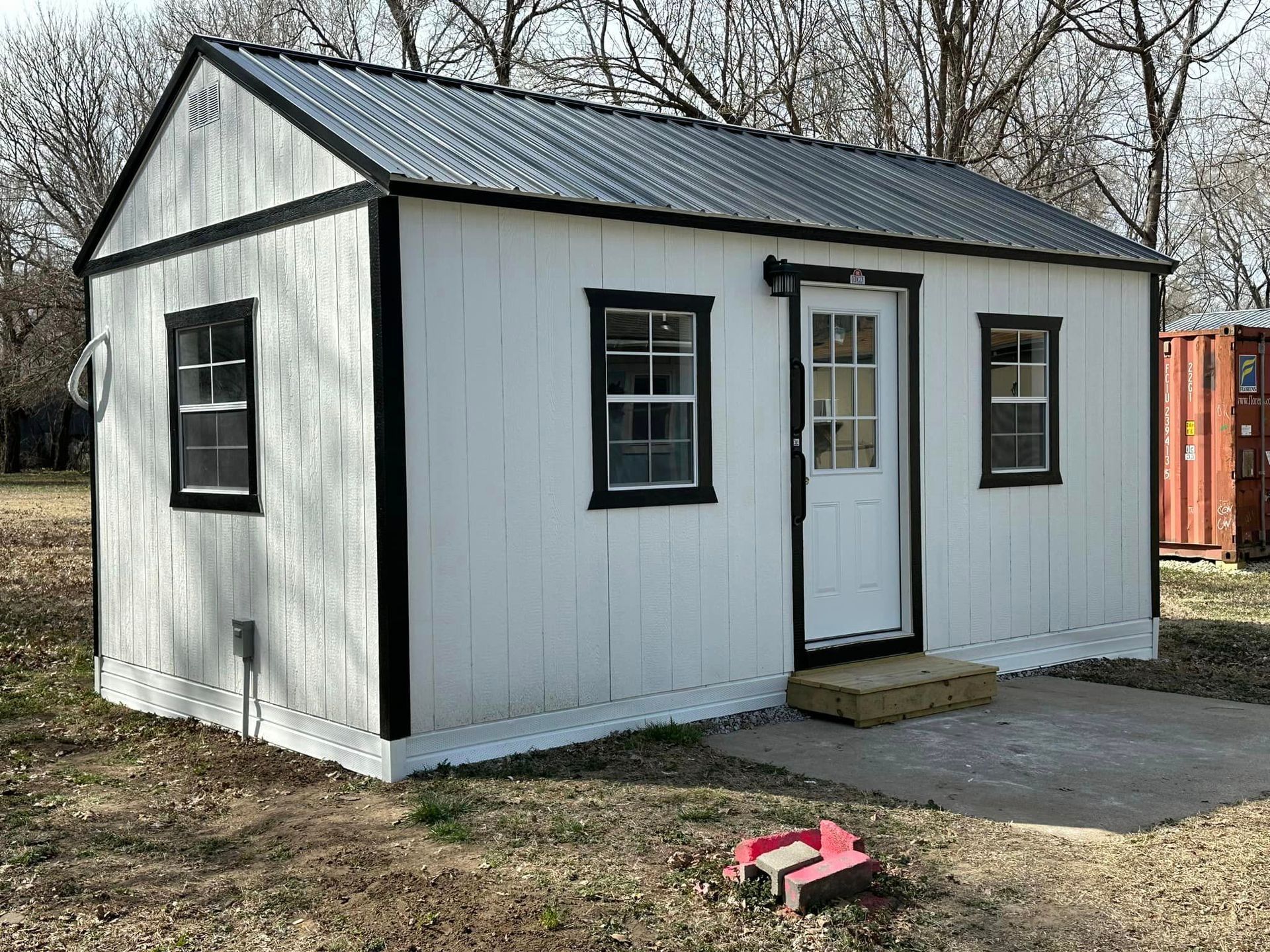 A small white shed with a black roof and windows is sitting in the middle of a field.