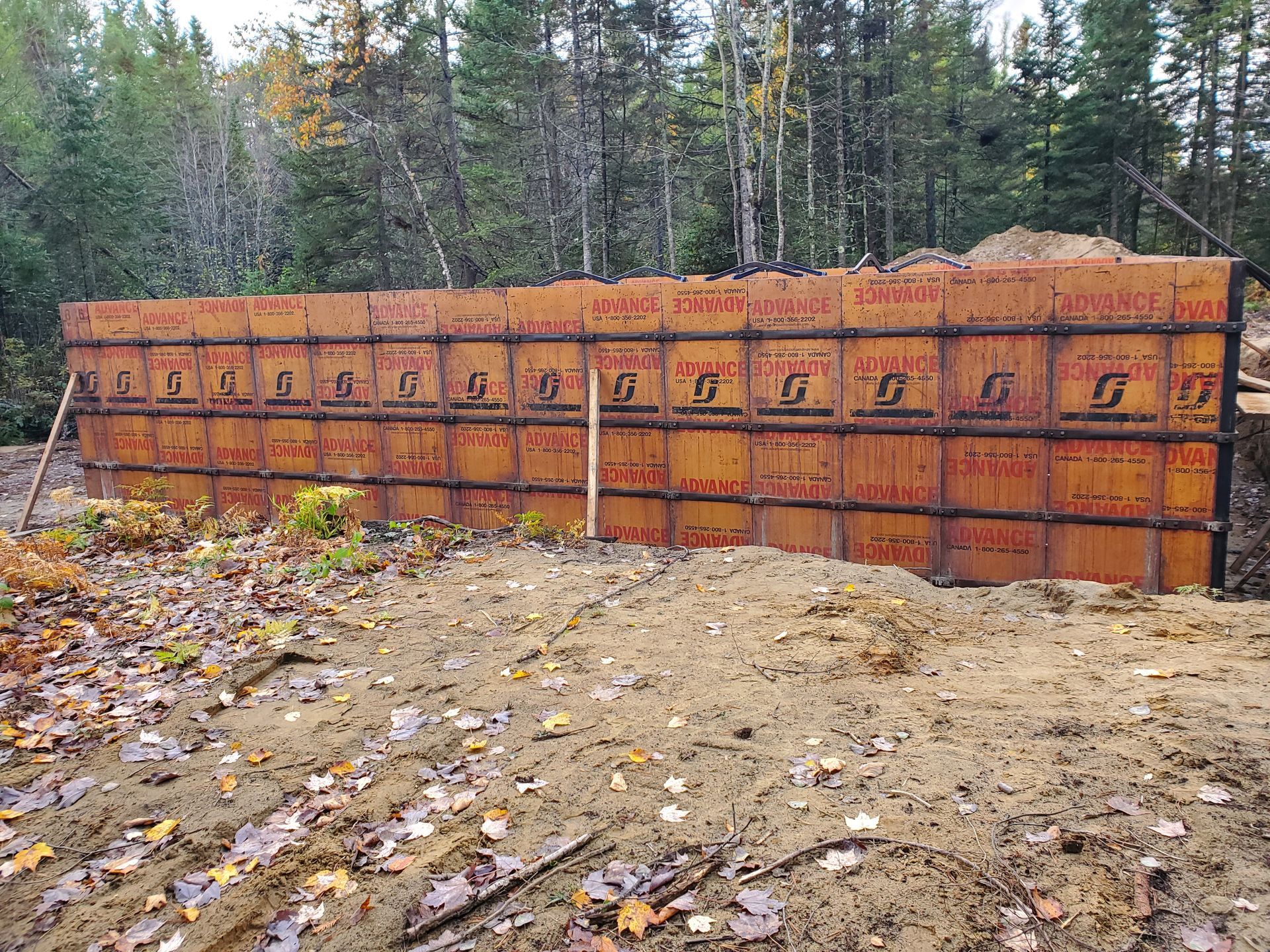Chantier de construction avec un grand mur de coffrage brun ; arrière-plan forestier et sol boueux.