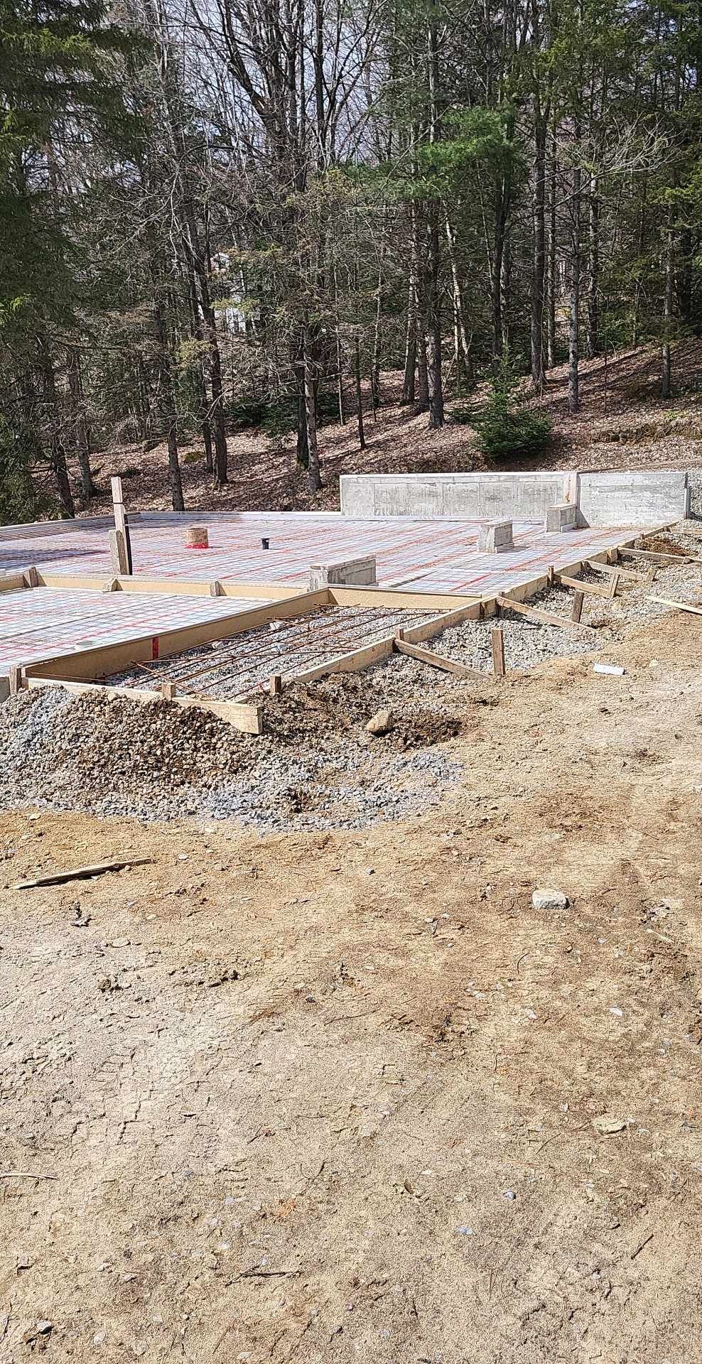 Chantier : Construction des fondations, gravier, charpentes en bois, forêt en arrière-plan.