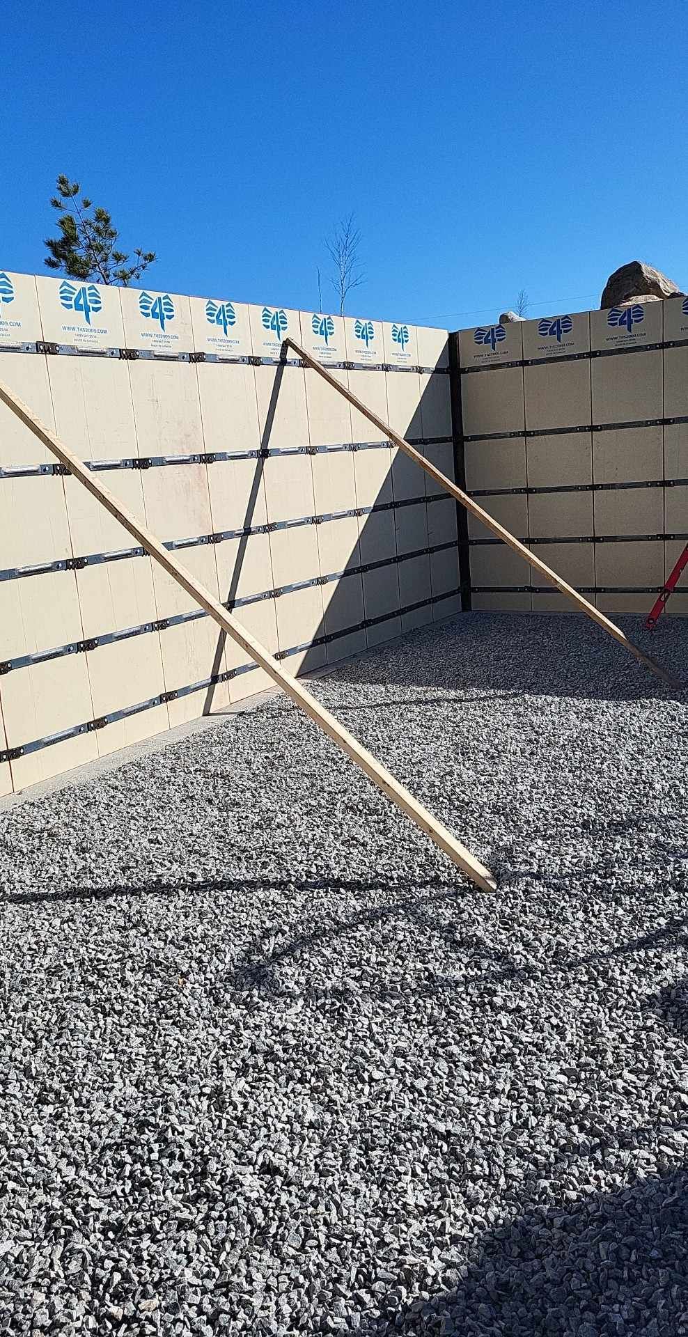 Chantier de construction avec coffrages en béton isolés, base de gravier et étais en bois contre le mur, sous un ciel bleu.