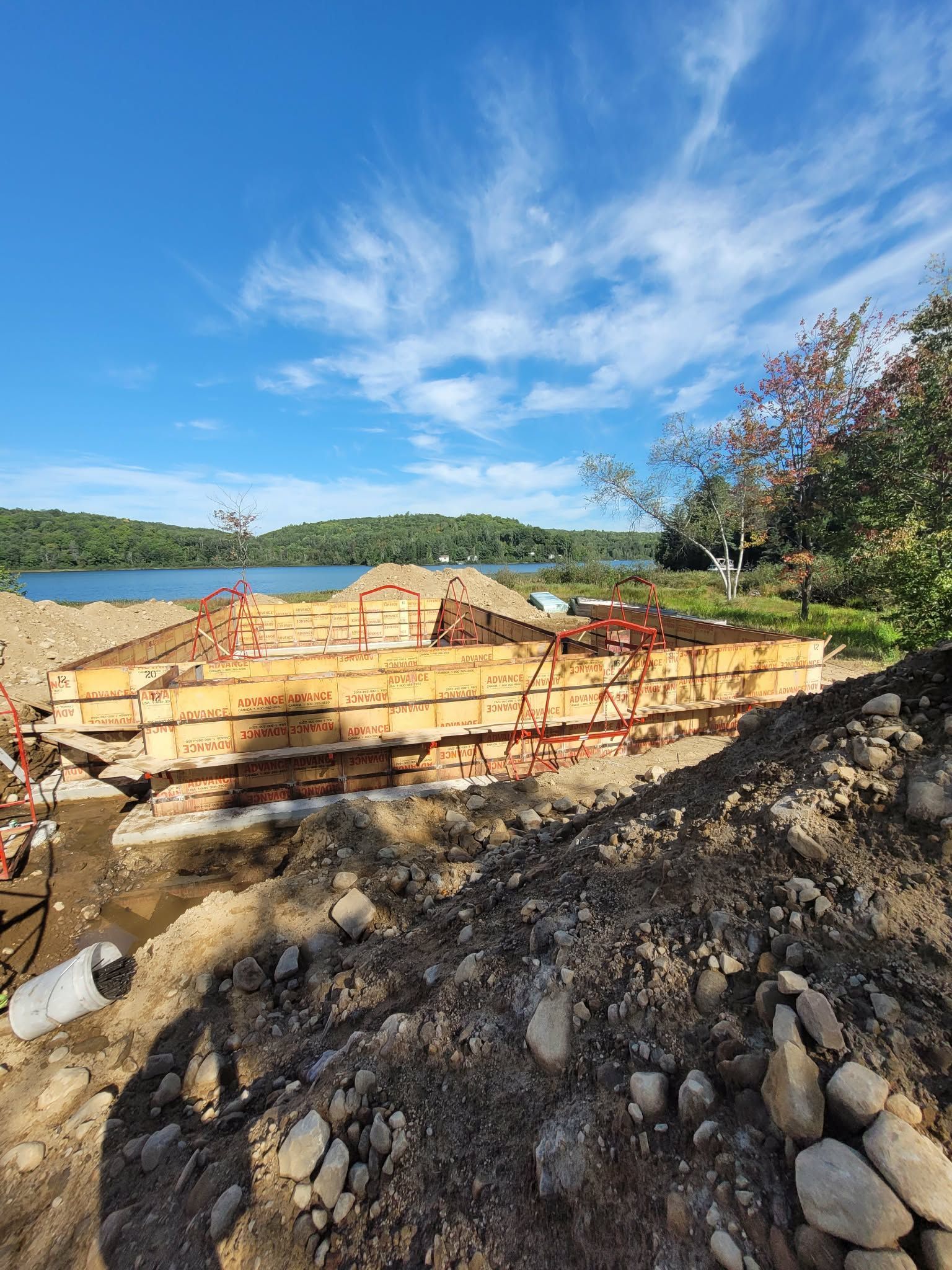Chantier de construction avec coffrages en bois pour fondations en béton, surplombant un lac sous un ciel bleu.
