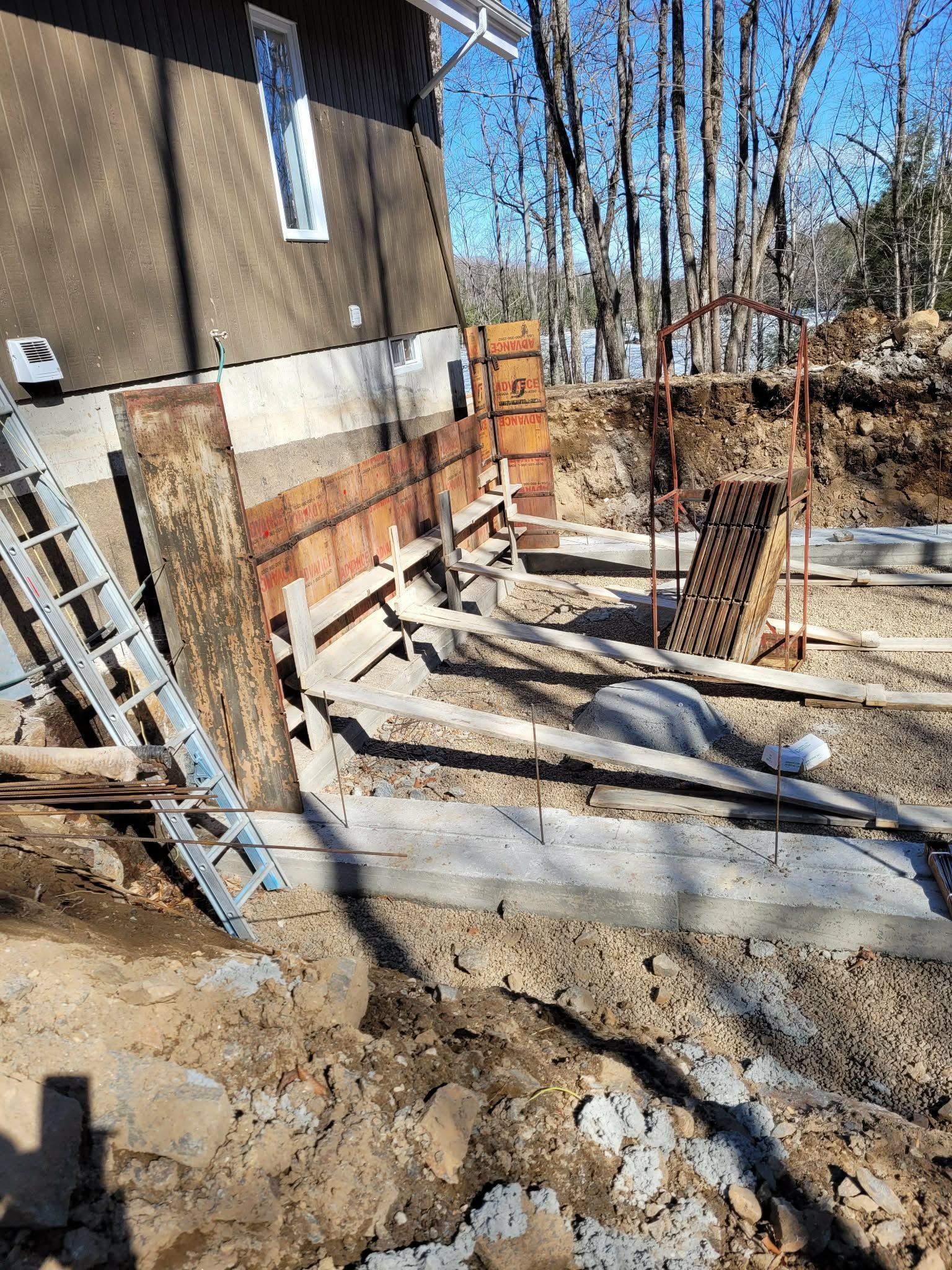 Chantier de construction avec coffrages en bois et barres d'armature près d'un bâtiment ; sol en terre battue.