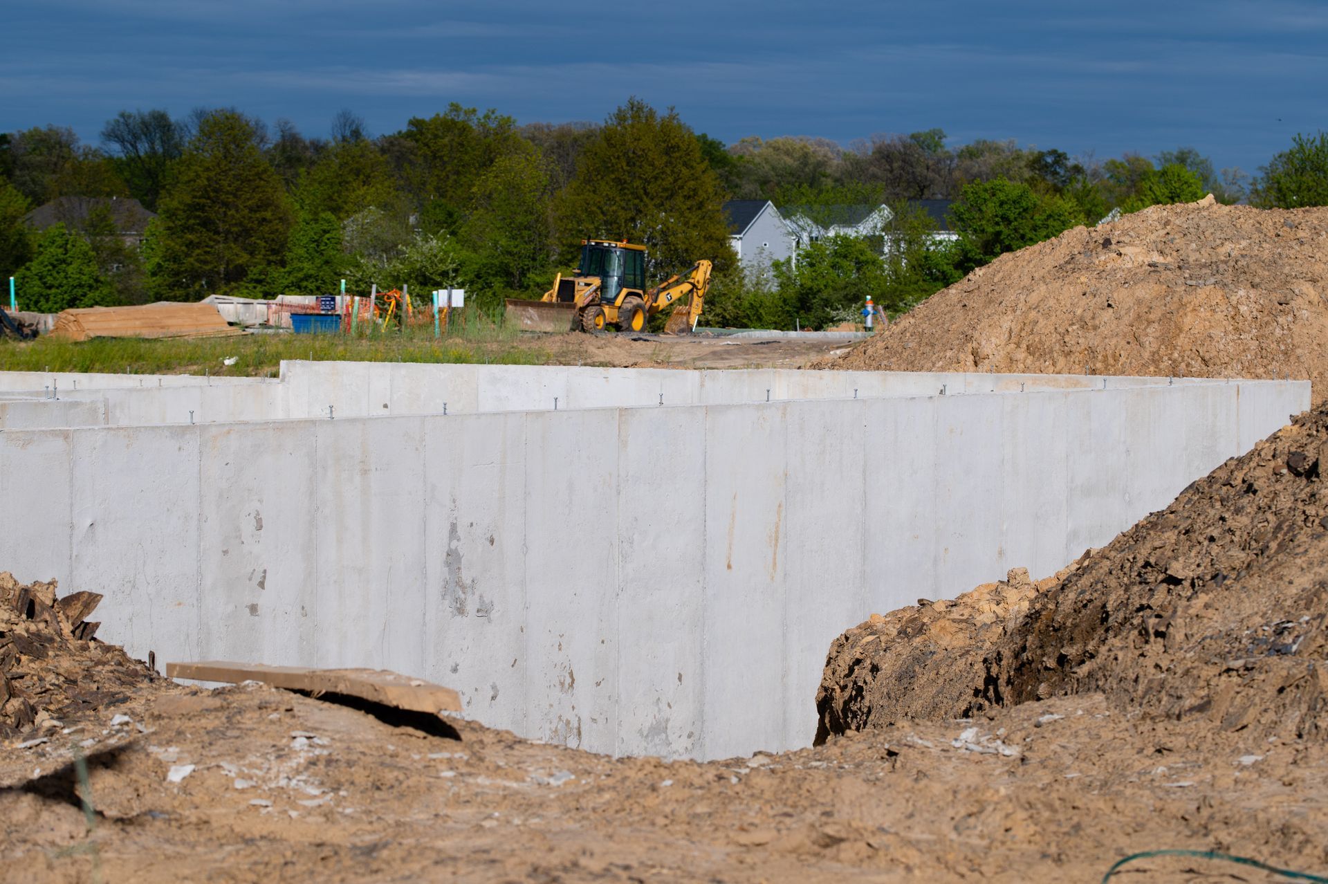 Mur de fondation en béton sur un chantier, tas de terre au premier plan, arbres et excavatrice au loin.