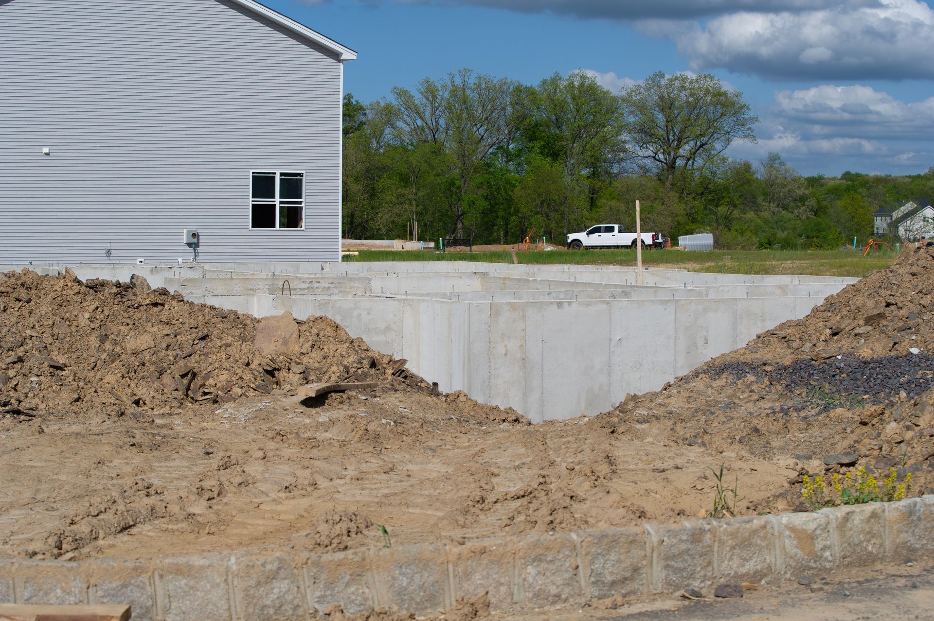 Fondations en béton en construction à côté d'un bâtiment à bardage métallique ; tas de terre au premier plan.