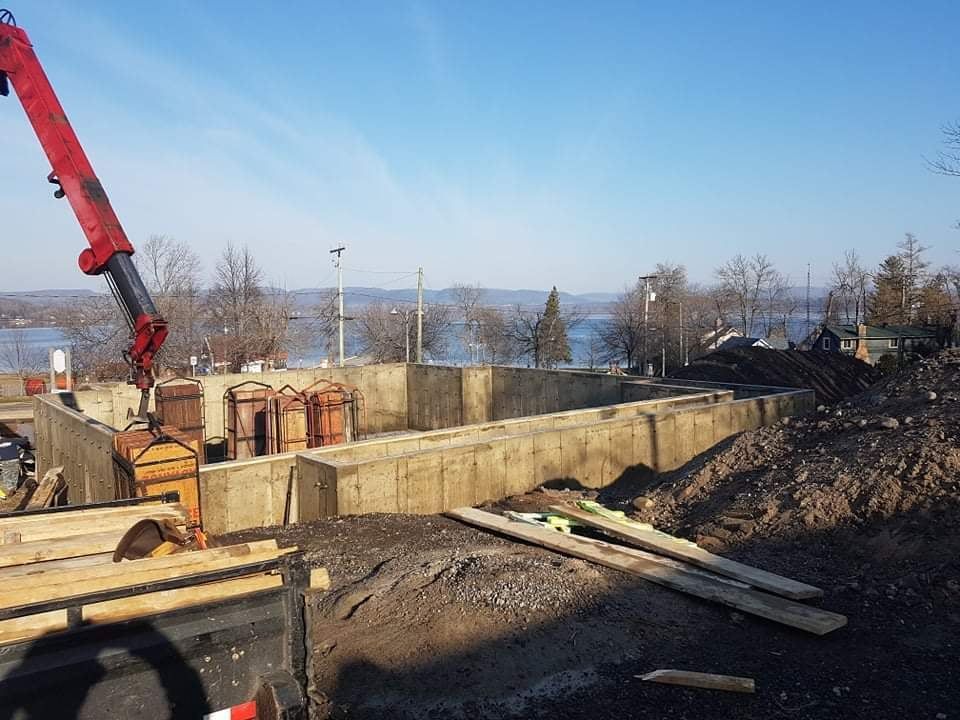 Chantier de construction avec murs de fondation en béton, bras de grue déployé et un lac en arrière-plan sous un ciel bleu.