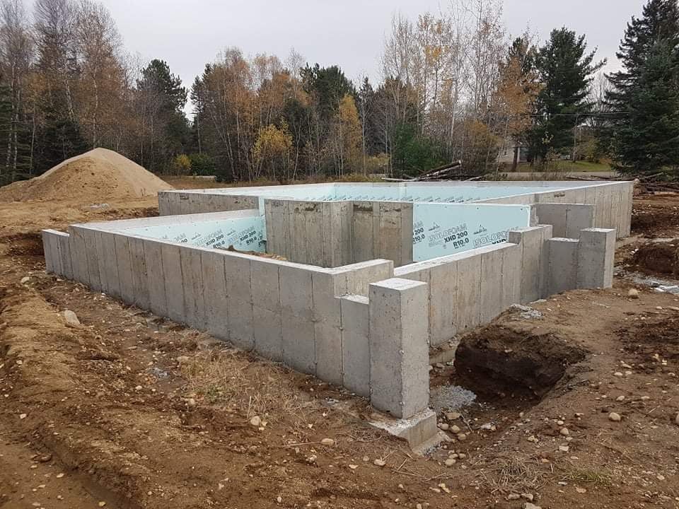 Fondations en béton d'un bâtiment en construction, entourées de terre et d'un tas de sable, avec des arbres en arrière-plan.