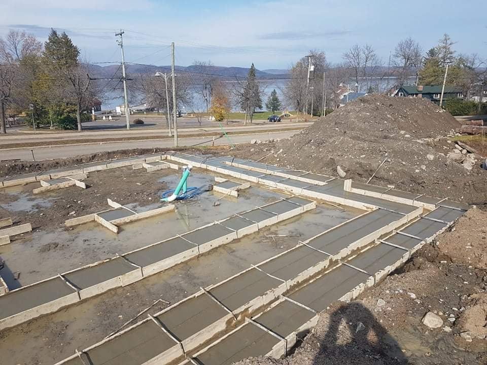 Fondations en béton en cours de construction, avec coffrages et béton frais. Un tas de terre se trouve à droite. Lac en arrière-plan.