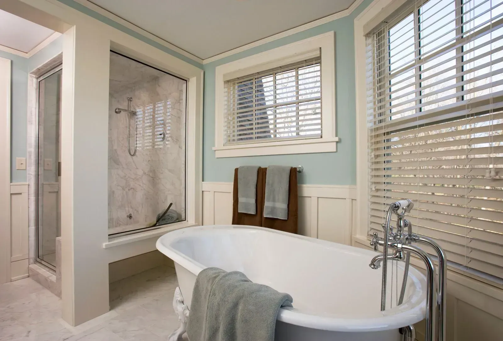 Bright bathroom with a freestanding tub, shower, and white shutters in soft gray and mint tones