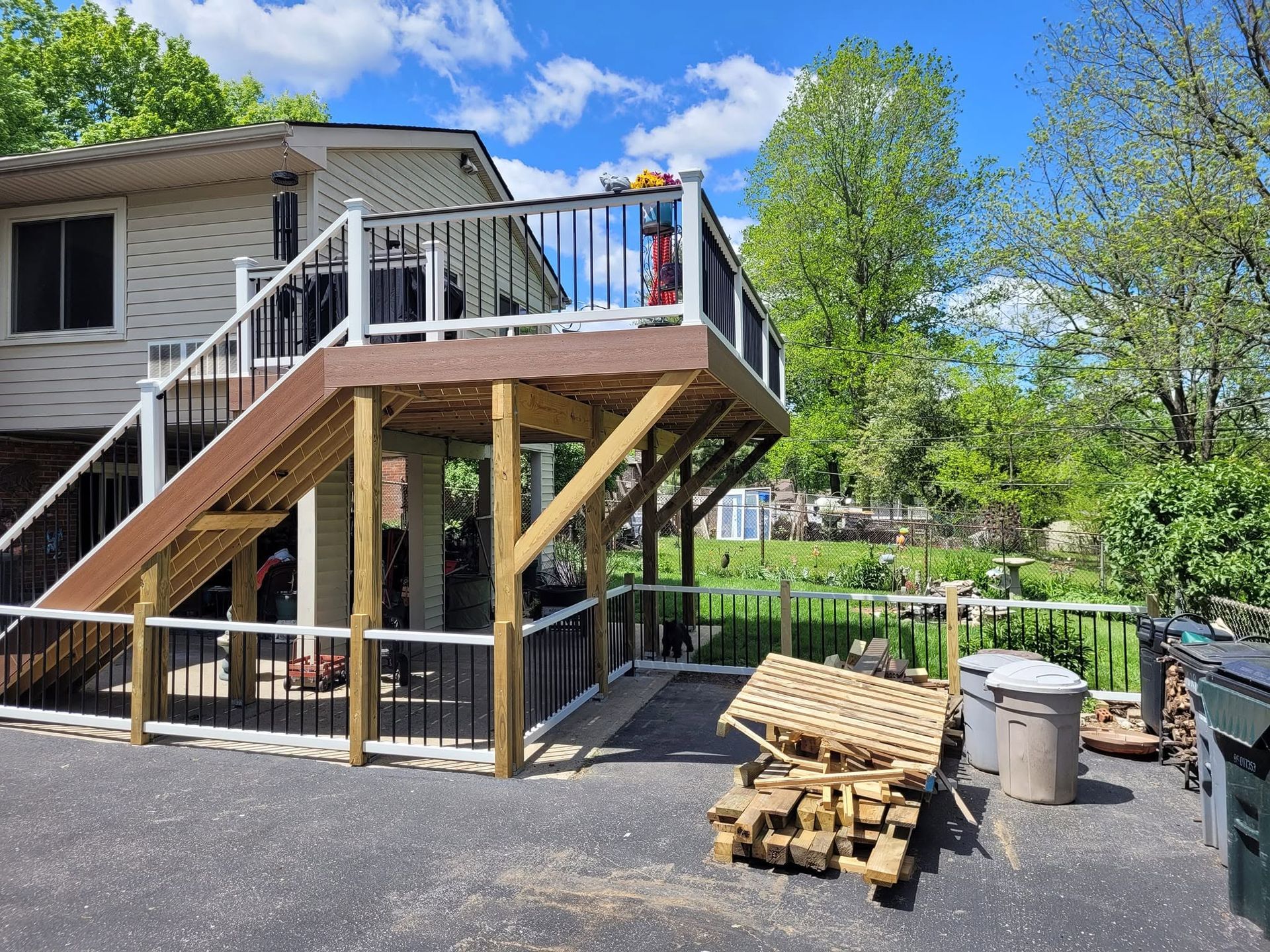 Deck stairs beside a house with lumber stacked on a driveway under a bright blue sky