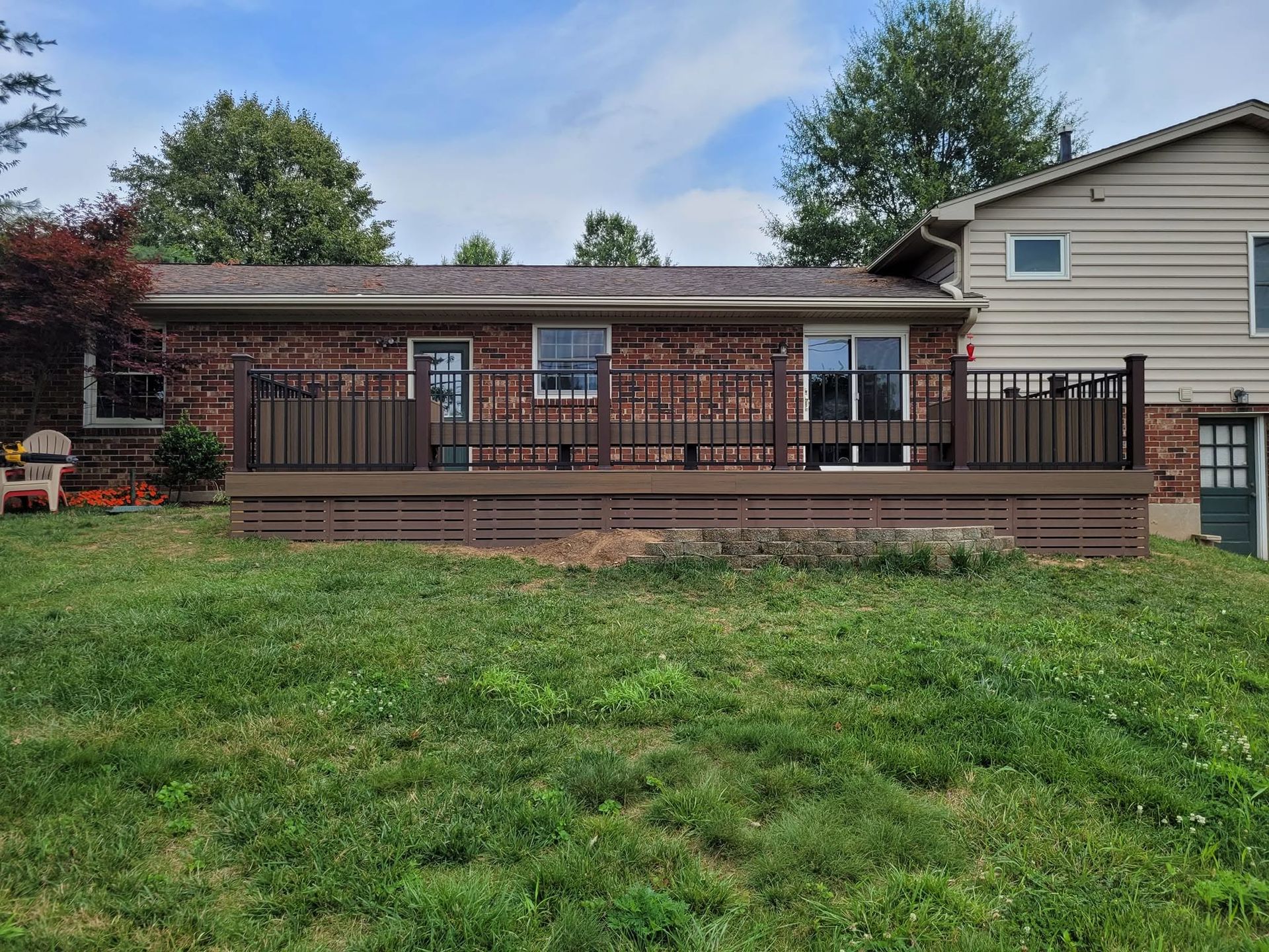 Backyard deck with black railing in front of a house, bordered by green lawn and trees.