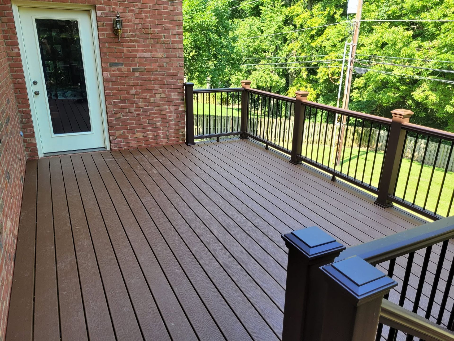 Wooden deck with black railing beside a brick house and door, overlooking green trees