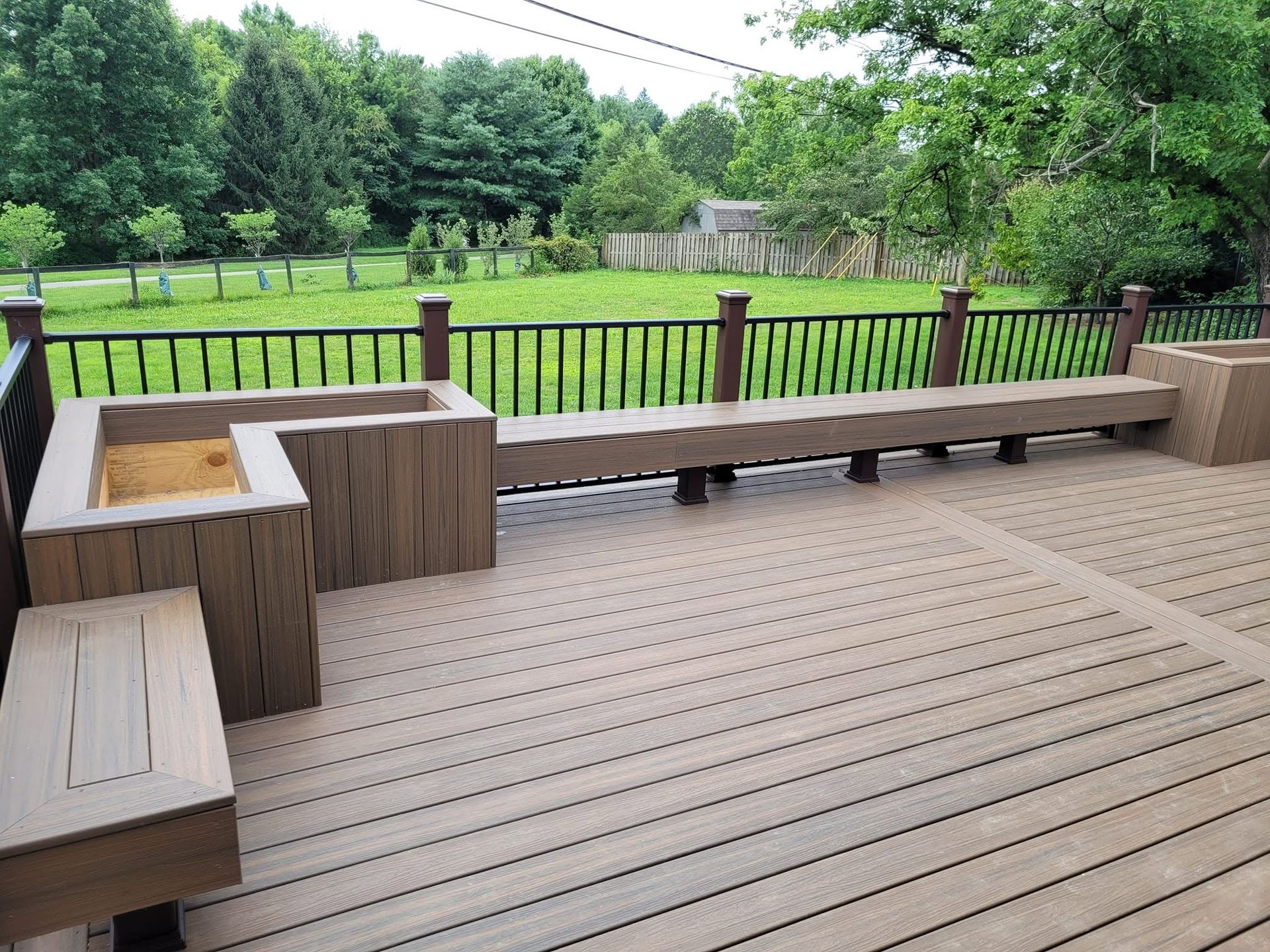 Wooden deck with built-in benches and a planter, overlooking a green backyard with trees and a fence.