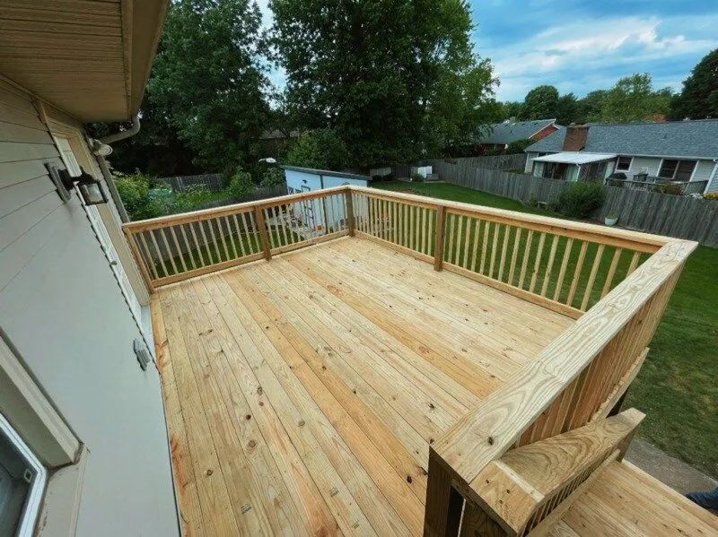 Wooden deck with railings, attached to a light-colored house. Green grass and trees are in the background.