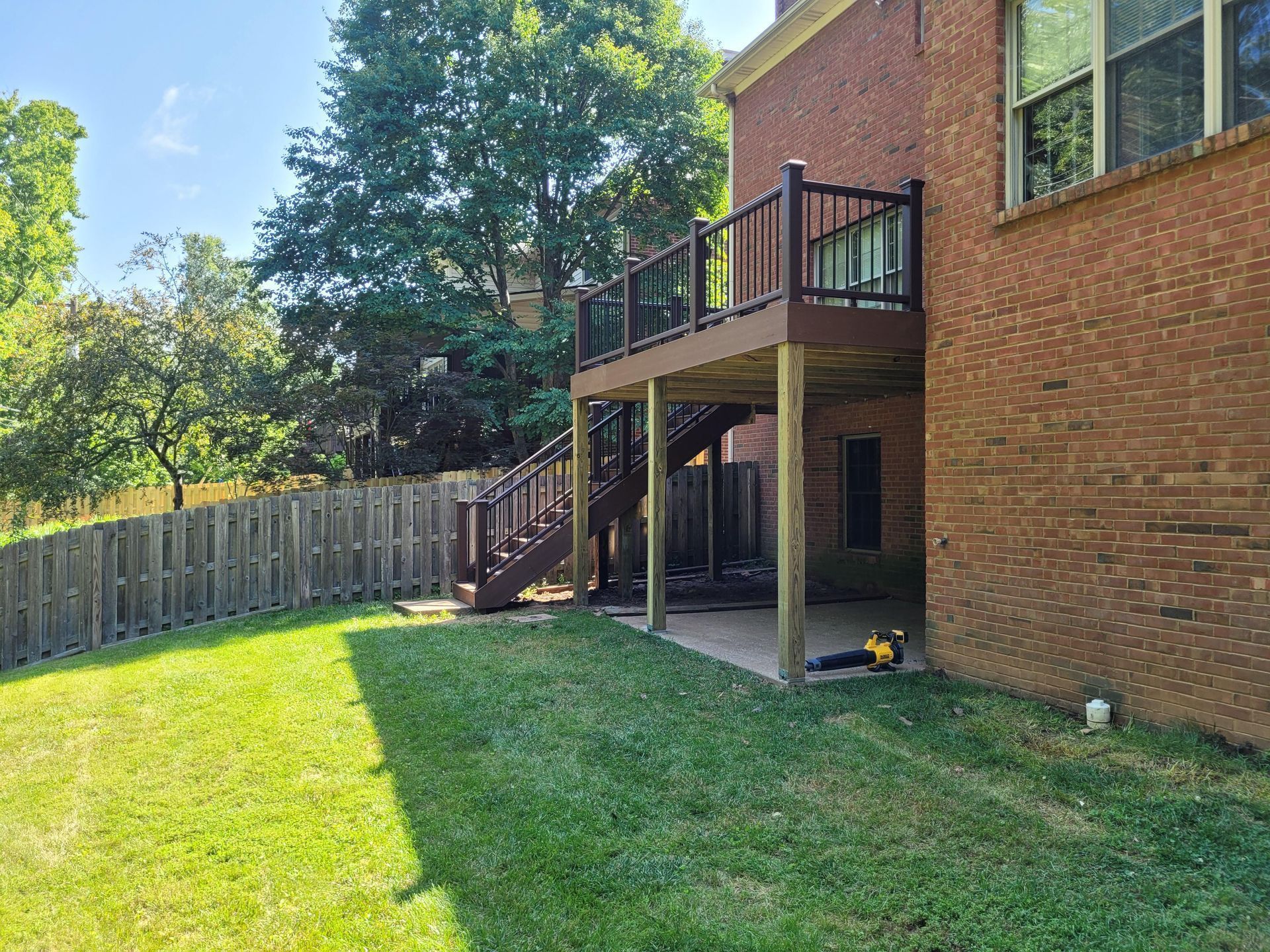 Backyard with green lawn, wooden fence, trees, and a brick house with a raised deck and stairs