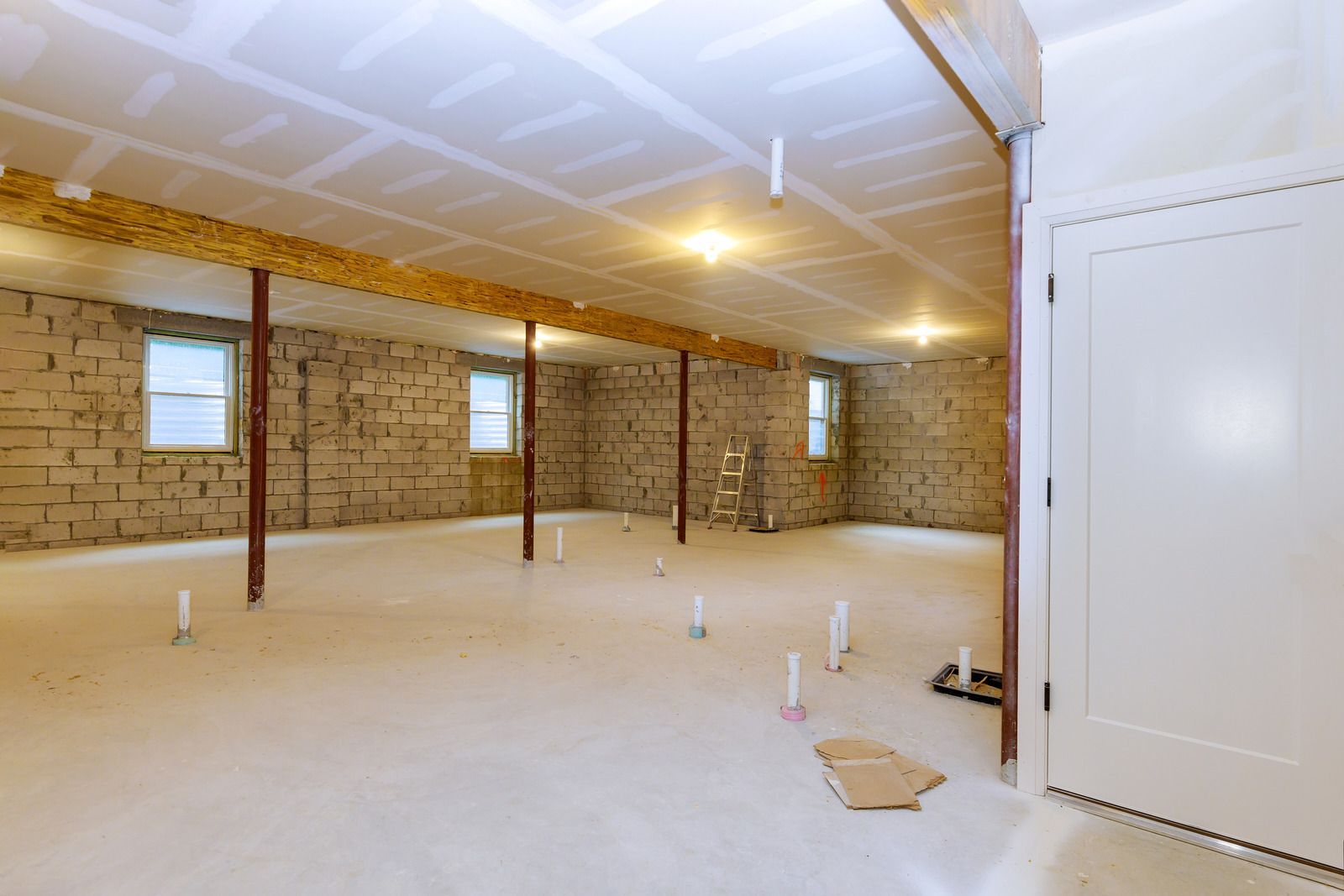 Basement under construction: exposed block walls, new drywall ceiling, pipes, support beams, and a white door.