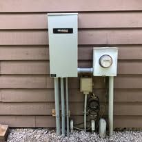 A electrical box is sitting on the side of a house next to a meter.