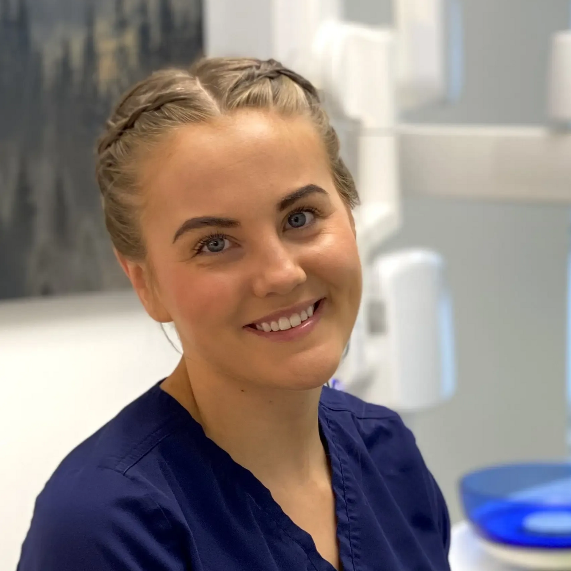 Smilende person med flettet hår på et tannlegekontor, iført blå skrubber.