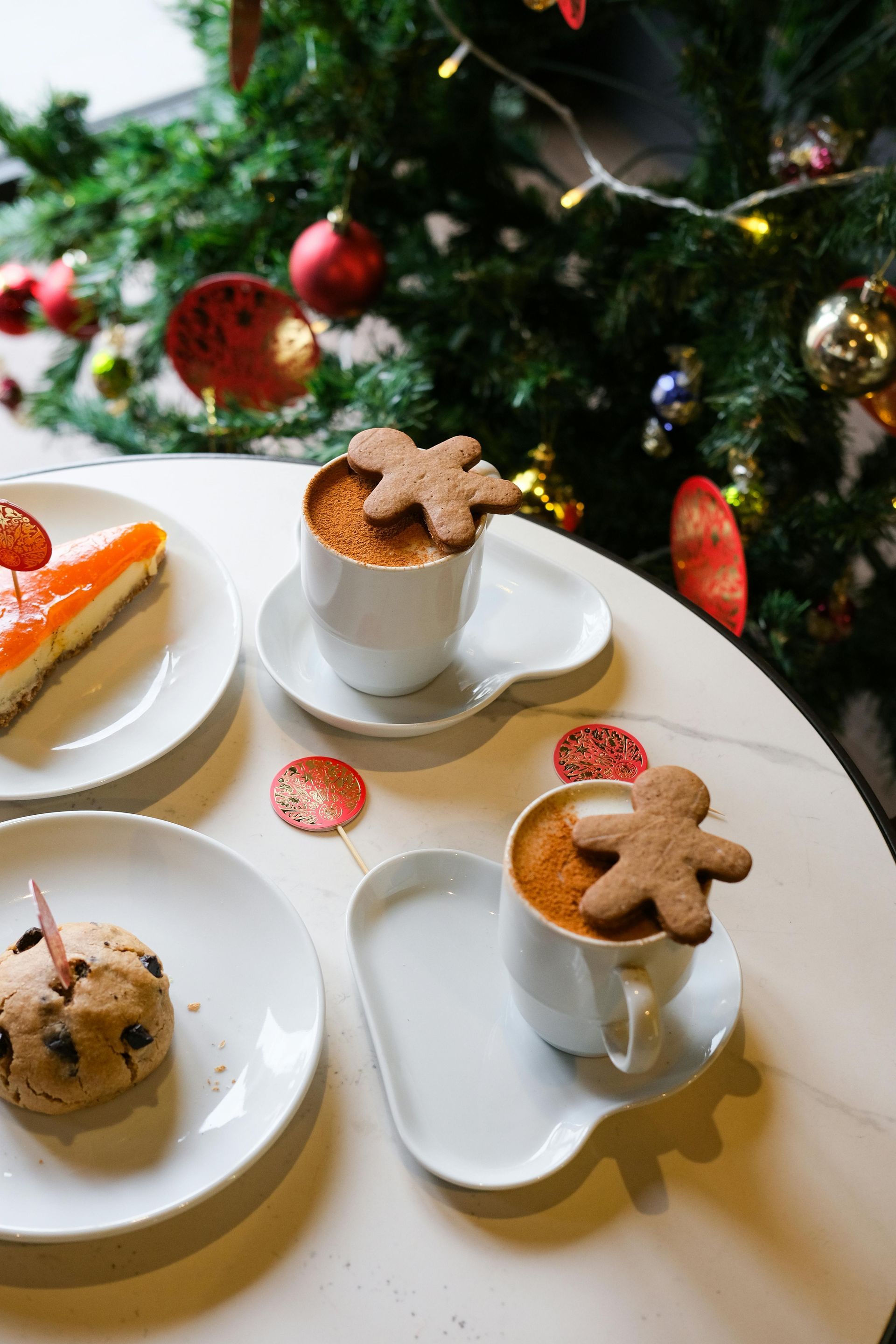 Two coffees with gingerbread men, cookies, and cake on a table near a decorated Christmas tree.