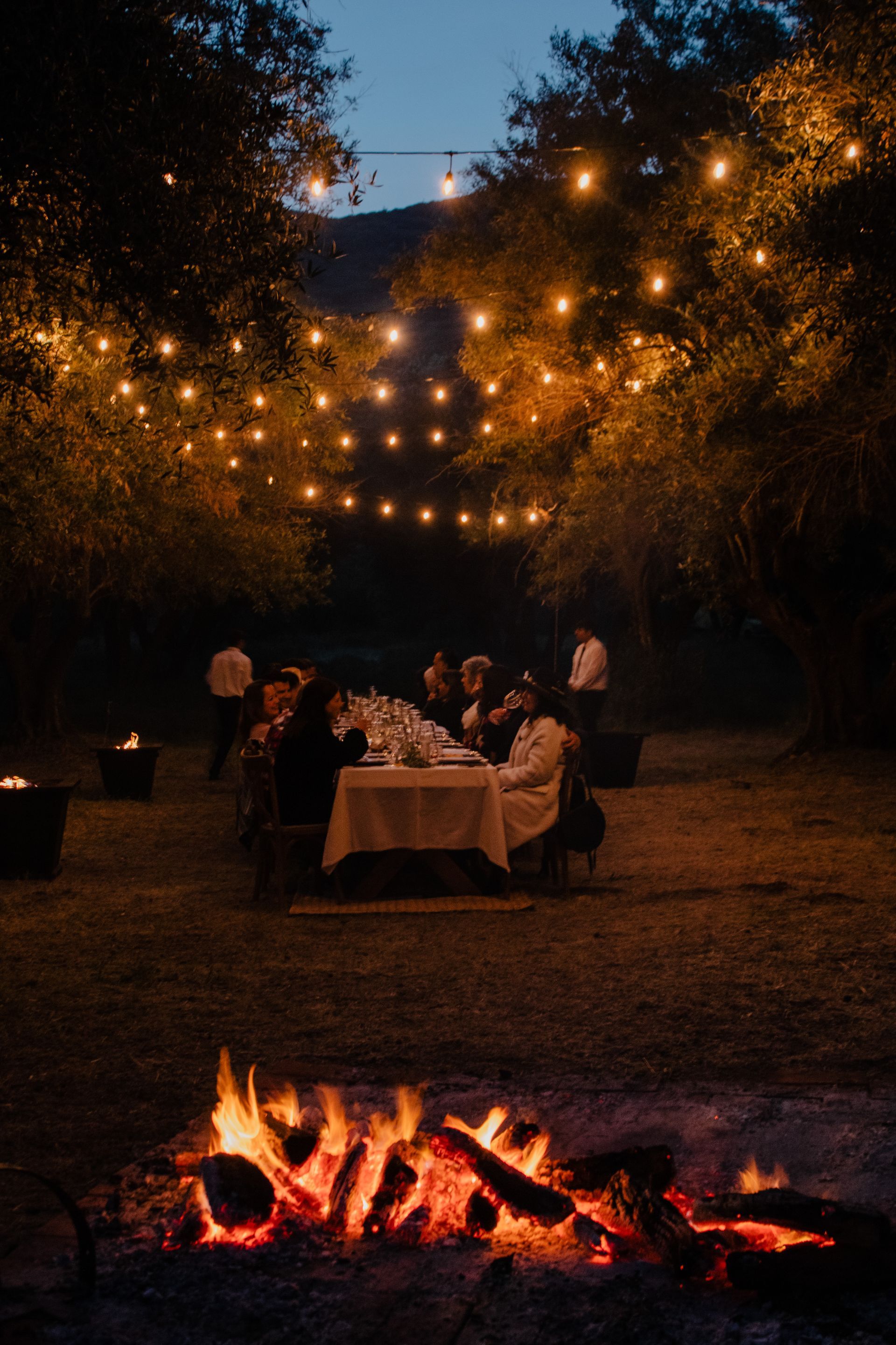 Cena al aire libre bajo guirnaldas de luces con una hoguera encendida en primer plano.