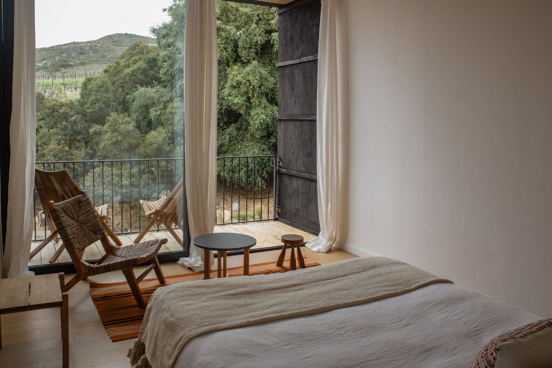 Bedroom with balcony overlooking a lush green landscape; two chairs, rug, and small table.