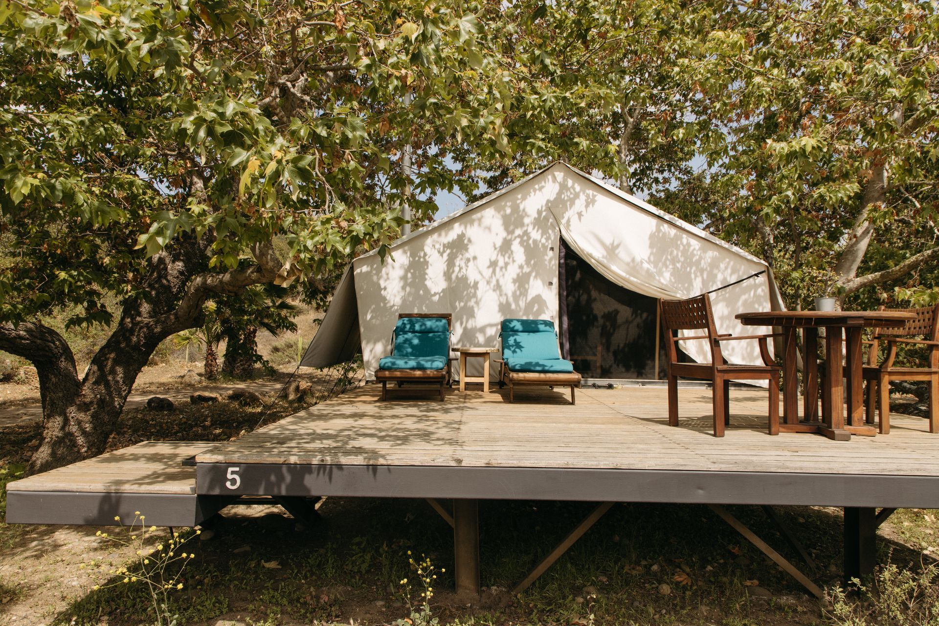 Tent on wooden platform with chairs and table, under trees.