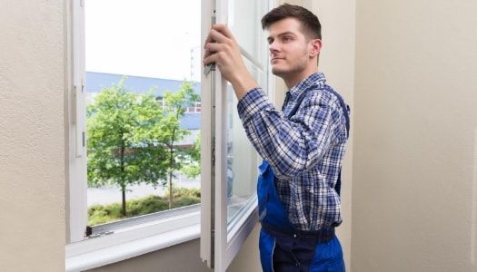 Window installer wearing a checkered blue shirt and blue overalls installing a new replacement window in the home.