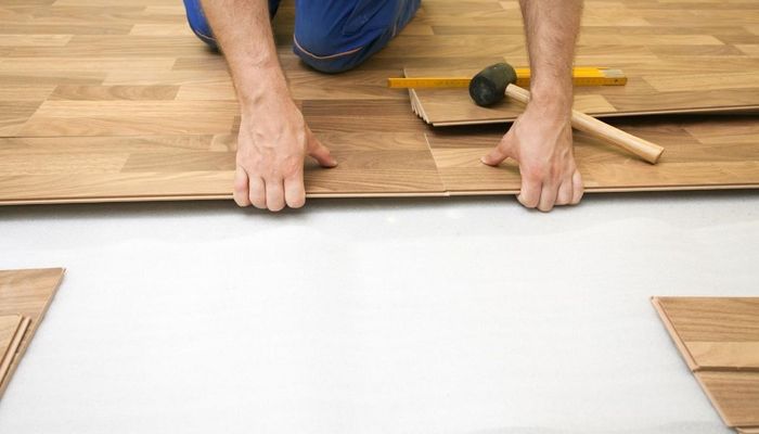 Burnaby Handyman installing wood laminate flooring with a square and a black mallet. The picture shows the man on his knees clicking the pieces into place.