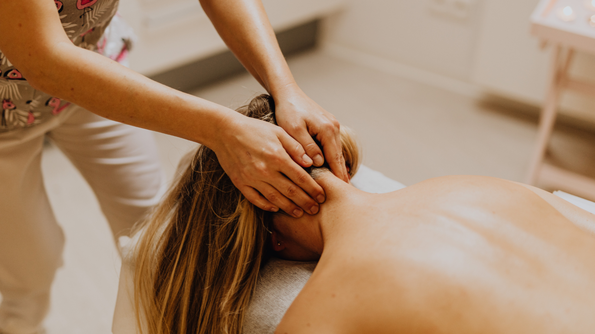 Hands massaging a person's neck and head. The person is lying face down on a massage table in a spa setting.