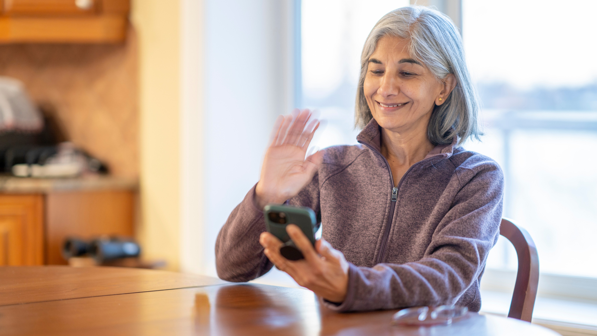 Woman with gray hair waves while holding a phone, sitting at a table in a kitchen.
