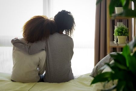 Two people sitting on a bed, leaning together by a bright window, with a plant on a shelf nearby