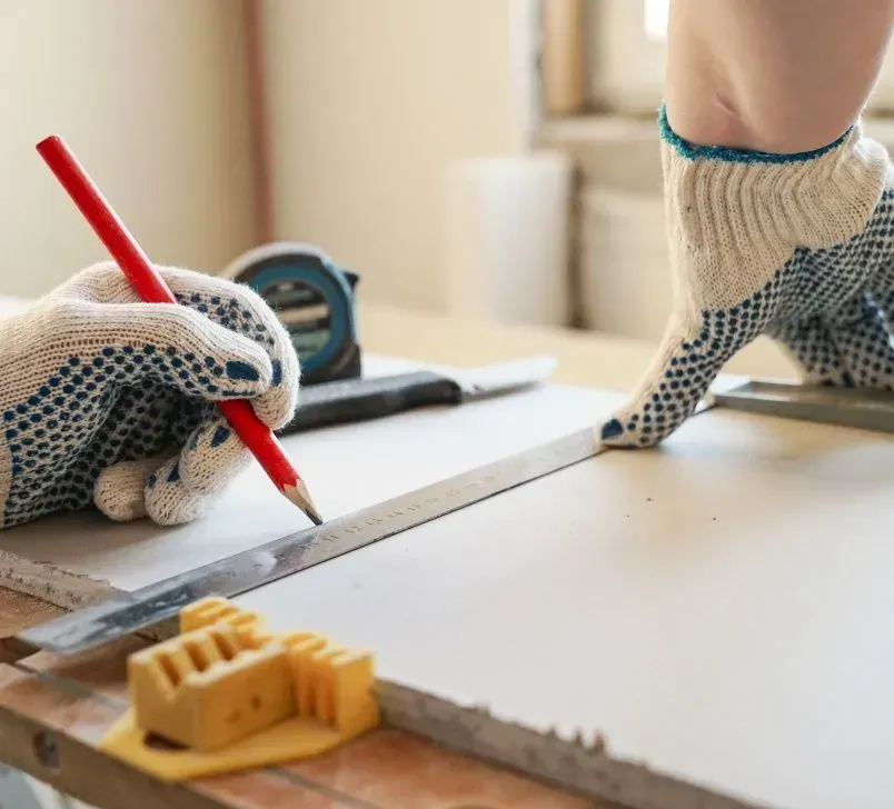 Person wearing work gloves using a pencil and ruler to measure and mark a white surface.