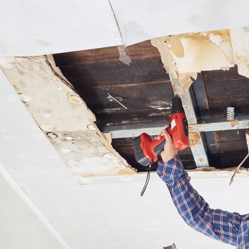 Person using a red drill to repair a damaged ceiling with exposed wood framing.